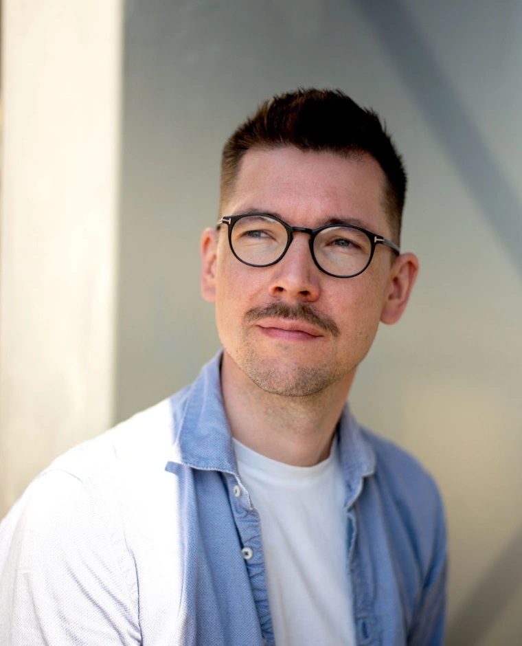 Eine Person trägt ein hellblaues Hemd über einem weißen T-Shirt. Der Hintergrund ist unscharf und grau.