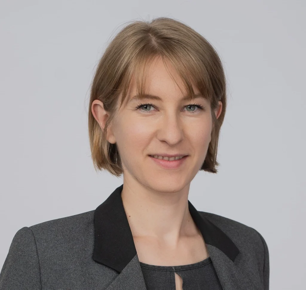 Person in einem grauen Blazer mit schwarzem Kragen und einem grauen Oberteil darunter. Hellbraunes, schulterlanges Haar. Hintergrund ist einfarbig hellgrau.