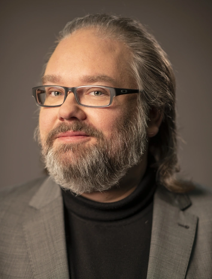 Eine Person in einem grauen Anzug und einem schwarzen Rollkragenpullover. Die Person hat grau meliertes, schulterlanges Haar und trägt eine Brille. Der Hintergrund ist einfarbig und dezent gehalten.