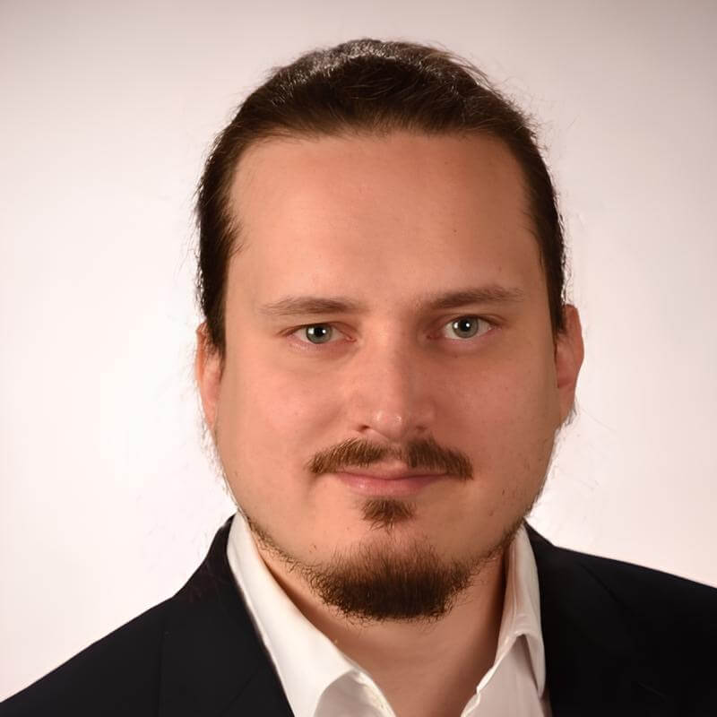 Professionelles Porträtfoto eines Mannes mit dunkelbraunem, nach hinten gekämmtem Haar und einem Kinnbart. Er trägt ein weißes Hemd mit offenem Kragen und ein dunkles Sakko. Der Hintergrund ist hell und neutral gehalten.