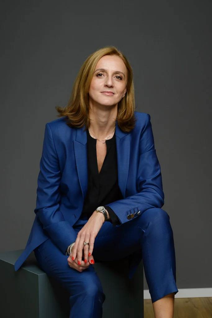 Professionelles Portraetfoto von Rechtsanwaeltin Dorela Kress. Sie sitzt auf einem grauen Wuerfel vor einem dunkelgrauen Hintergrund. Sie traegt einen blauen Hosenanzug ueber einer schwarzen Bluse, eine Armbanduhr und Schmuck an den Haenden sowie rot lackierte Fingernaegel. Sie hat schulterlanges blondes Haar und blickt freundlich und selbstbewusst in die Kamera. Der Boden ist aus hellem Holz.