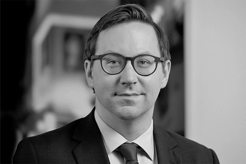 Schwarz-Weiss-Portraetfoto von Dr. Christopher Unseld. Er traegt eine Brille mit dunklem Gestell, einen dunklen Anzug mit weissem Hemd und dunkler Krawatte. Er hat kurzes seitlich gescheiteltes Haar und blickt freundlich in die Kamera. Der Hintergrund ist unscharf.